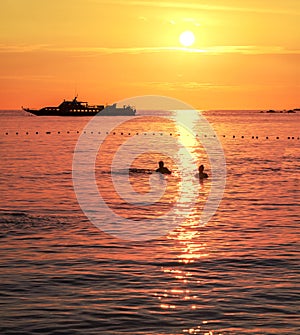 Silhouettes of a swimming couple and ship at sunset.