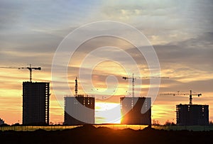 Silhouettes of tower cranes constructing a new residential building at a construction site against sunset background.