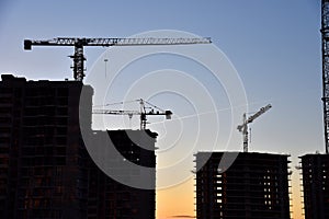 Silhouettes of tower cranes constructing a new residential building at a construction site on sunset background.