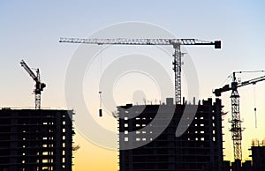 Silhouettes of tower cranes constructing a new residential building at a construction site on sunset background. Crane lifting a