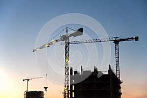 Silhouettes of tower cranes constructing a new residential building at a construction site on sunset background. Crane lifting a