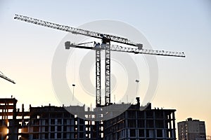 Silhouettes of tower cranes constructing a new residential building at a construction site on sunset background. Crane lifting a