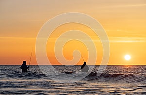 The silhouettes of two anglers in waterproof trousers fishing in front of a dreamy orange sunset in the Baltic Sea.