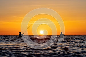 The silhouettes of two anglers in waterproof trousers fishing in front of a dreamy orange sunset in the Baltic Sea.