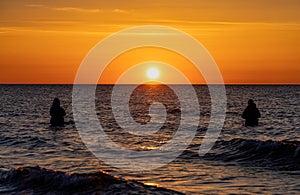 The silhouettes of two anglers in waterproof trousers fishing in front of a dreamy orange sunset in the Baltic Sea.