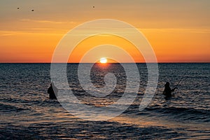 The silhouettes of two anglers in waterproof trousers fishing in front of a dreamy orange sunset in the Baltic Sea.