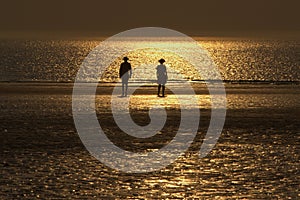 Silhouettes of two people walking on the north sea beach with reflection of the setting sun in the water and sand