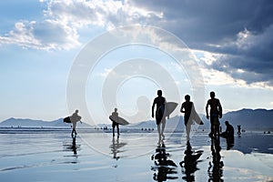 Silhouettes of unrecognizable surfers carrying their surfboard on sunset beach, with a cloud and mirroring in the water