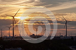 Silhouettes of wind turbines under a cloudy sky at sunset with a shining golden sun peeking through