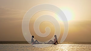 Silhouettes, woman and teen girl, mom and daughter, sit on edge of outdoor infinity pool with panoramic sea view and
