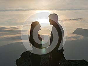 Silhouettes of young couple standing on a mountain and looking to each other on beautiful sunset background. Love of guy