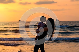 Silhouettes young mother with daughter playing and kissing on the beach at sunset evening sky background. Happy family.