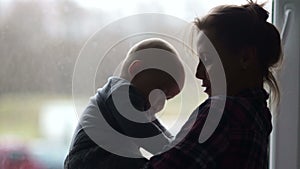 Silhouettes of a young mother and a six-month old child against the background of a window. Housewife on maternity leave