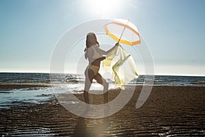 Silhoutte of a pretty senior Asian female in a swimming suit standing and hold umbrella and flaunt yellow fabric on the beach.