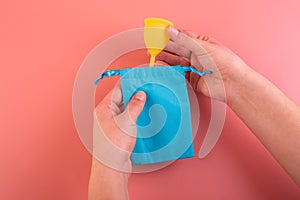 Silicone yellow menstrual cup with blue pouch in female hand. Women's health and alternative hygiene on a pink background