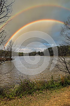 A silky brown still lake surrounded by lush green trees and blue sky and clouds reflecting off the water and a rainbow in the sky