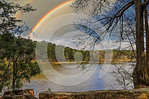 A silky brown still lake surrounded by lush green trees and blue sky and clouds reflecting off the water and a rainbow in the sky