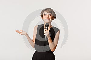 Silly cute brunette girl looking indecisive up, holding microphone, singing song, standing over white background