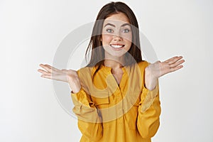 Silly and cute young woman shrugging with hands spread sideways and awkward smile, saying sorry as being clueless