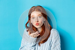 Silly redhead woman in sweater, blowing air kiss at camera with puckered lips, standing against blue background