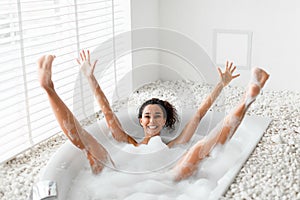 Silly young woman having fun in bubble bath, raising her arms and legs up, being silly and playful at home