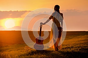 siluette of woman wearing a bohemian style with guitar on a field at warm light of sunset