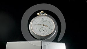 Silver antique pocket watch on a stand rotating on a black isolated background. Round gray retro clock with a white dial