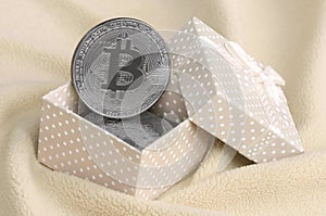 The silver bitcoin lies in a small orange gift box with a small bow on a blanket made of soft and fluffy light orange fleece