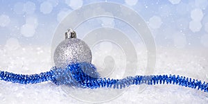 Silver Christmas ball with blue festive tinsel in the snow on an abstract bokeh background