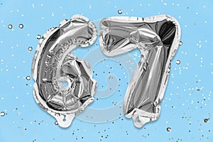 Silver foil balloon number, digit sixty seven on a blue background with sequins. Birthday card with inscription 67