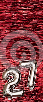 Silver foil balloon number number 27 on a background of red tinsel decoration. Birthday greeting card, inscription