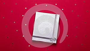 Silver gift box on deep red background with hearts confetti, copy space, top view, flat lay. Background for Valentine`s Day,