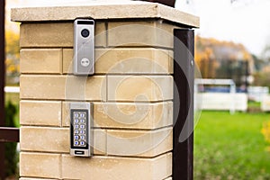 Silver intercom call panel with blue number buttons and a video camera on a brick beige fence post of a private house