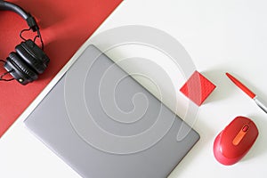Silver laptop, red computer mouse, wireless headphones, pen and red box on red and white background. Concept of a modern workplace