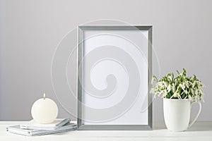 Silver photo frame in modern scandinavian style with bouquet snowdrop flowers in the ceramic cup and burning candle on wooden