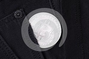 A silver physical Bitcoin coin sticks out of the pocket of black jeans close-up.