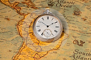 Silver retro pocket watch lying on a paper old world map. Antique round clock with a dial and arrows on a geographical