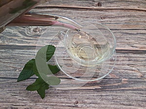 Silver teapot pouring peppermint tea (Mentha spicata) into a clear glass cup on a wooden table.