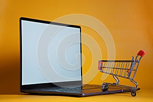 Silver ultrabook and supermarket trolley on a yellow background. The concept of shopping on modern online internet commerce sites