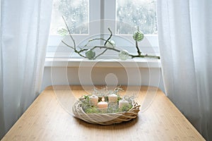 Simple advent wreath with moss and white candles, the first one is lit, on a table near the window on a snowy winter day, holiday