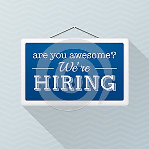 Simple blue sign with text `we`re hiring` hanging on a gray office wall. Human Resources, employment concept