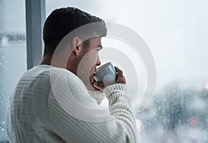 A simple break will bring you back to you. Shot of a young man having coffee and looking out of a window on a rainy day