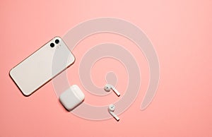 Simple flat lay photo on a pastel pink background with a white smartphone and wireless earphones with a charging case