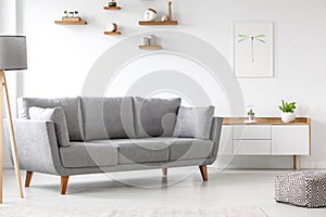 Simple, gray sofa standing next to a white cupboard in living room interior with decorations on wooden shelves. Real photo