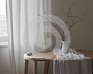 The simple interior of the living room - a round wooden table with a jug with branches and a chair. Minimalism style