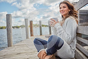 A simple life is its own reward. a beautiful young woman enjoying a warm beverage while relaxing on a bench at a lake.