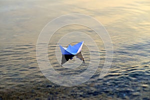 Simple small white origami paper boat floating quietly in blue clear river or sea water under bright summer sky.