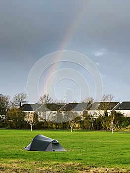 Simple tent on green grass in a park. Row of houses in the background. Rainbow in a blue sky. Homeless living close to dense