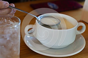 Simple white cream soup in Stainless spoon from a white ceramic cup with two handles on saucer and a glass of cool water beside.