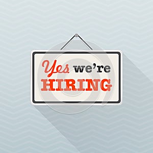 Simple white sign with text `Yes we`re hiring` hanging on a gray office wall. Human Resources, recruiting concept.